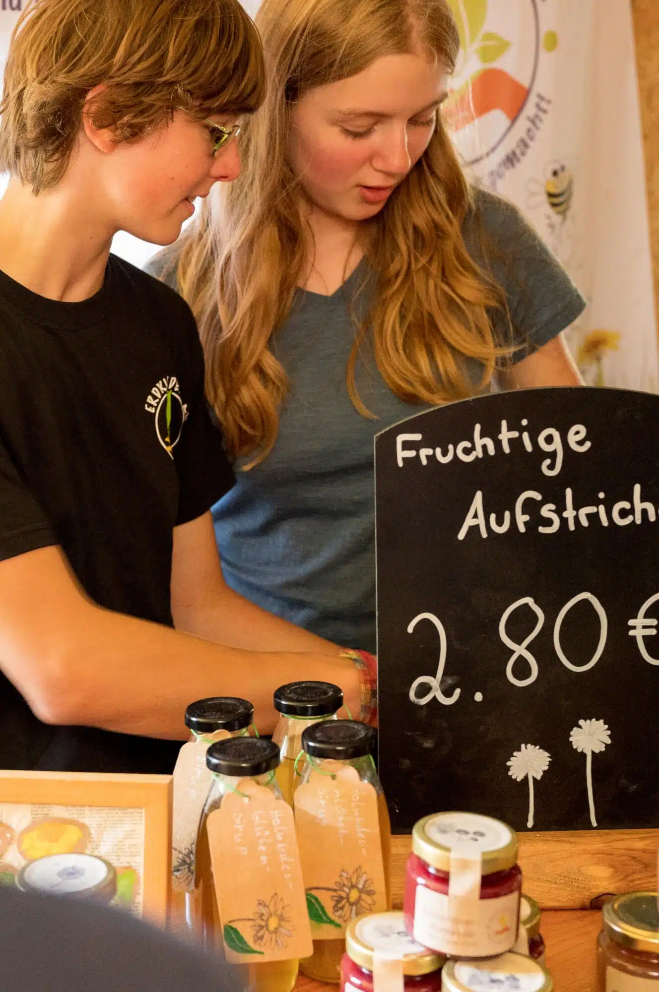 Two Erdkinderclan students behind their market stall, with bottles of elderflower syrup and a&nbsp;chalkboard reading Fruchtige Aufstriche, 2.80 euros.